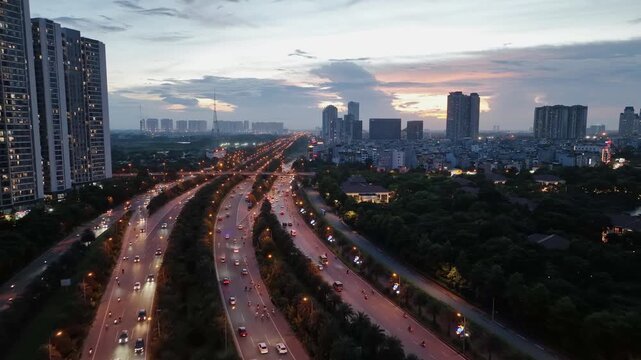 Drone view of curved boulevard in Hanoi with steady traffic, surrounding greenery, and residential towers under soft evening sky near sunset