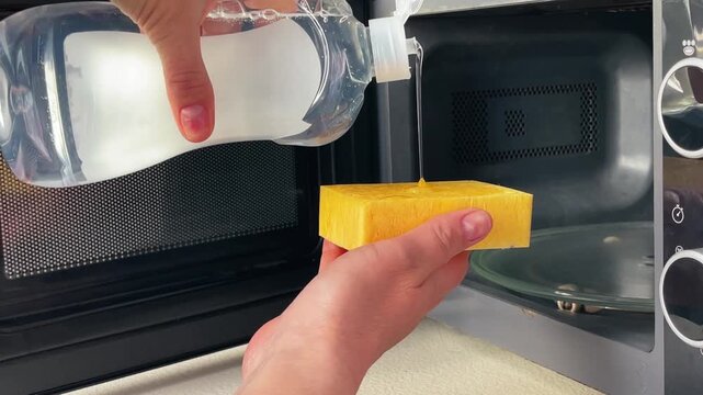 Close-up of pouring detergent liquid onto a yellow sponge in front of open microwave