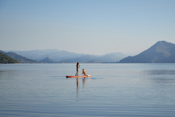 A woman stands with a Nova Scotia Duck Tolling Retriever on a board surrounded by peaceful water. The pair is paddling together against a mountainous backdrop. © Anna Averianova