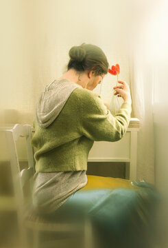 Woman sitting in front of a window in a meditative and romantic pose with a red tulip in her vase on a wooden shelf in the background VI