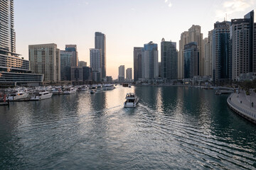 Obraz premium 12.12.2025 Dubai, UAE. Luxury yachts and boats moored in Dubai Marina at sunset. Stunning view of skyscrapers and modern architecture in UAE with golden sky reflection in the water. High-end lifestyle