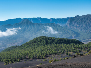 Fototapeta premium Solitary Pine on Volcanic Ridge View - Single pine tree on a black volcanic slope with a panoramic view of the vast mountain range and cloud formations under a clear blue sky.