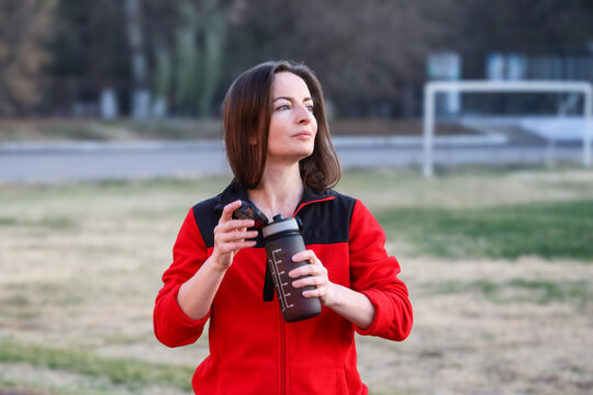 Fit mid adult brunette woman in her 40s drinking water from a sports bottle after outdoor stadium workout