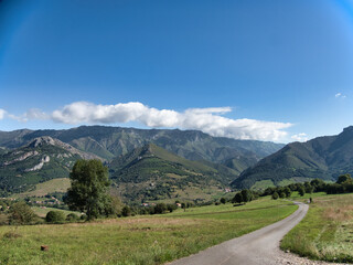 Fototapeta premium Winding Road Through Mountain Valley in Asturias - Scenic view of a paved road cutting through lush green meadows with rolling mountains under a bright blue sky.