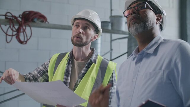 Foreman in hard hat and safety vest discussing architectural plan and construction site details with senior architect inside unfinished building