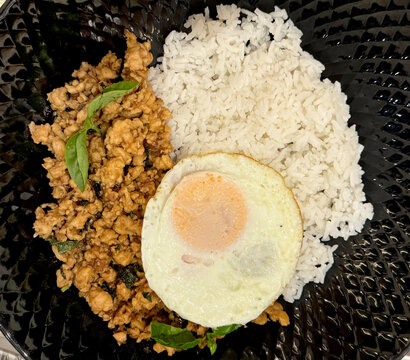 Thai herbs chicken basil with seasoned Thai rice. Served with sunny sided egg, and coriander leaves, inside a white plate, on a wooden rustic table.