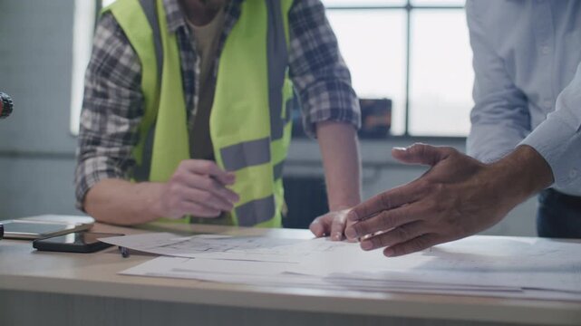 Close up view of foreman and architect holding pencils and discussing floor plan details at desk with tools and documents on construction site