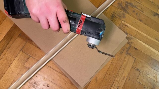 Hand holds a cordless power tool cutting cardboard on a wooden floor, close view of diy work in progress, practical home improvement mood