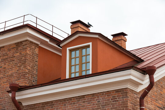 A red roof dormer with a multi-pane window and brick chimneys on a historic urban building, highlighting architectural textures, warm earth tones and classic exterior details against an overcast sky