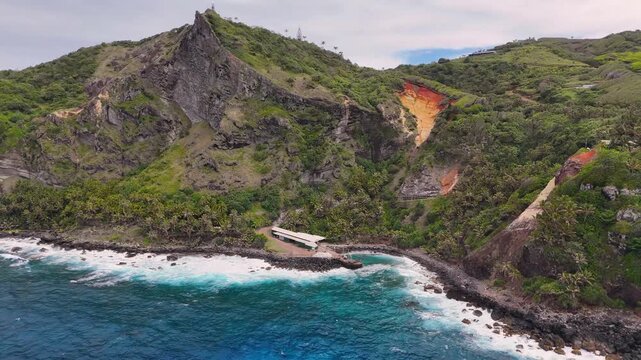 Aerial Drone footage of Pitcairn Islands Over Clear Blue Ocean. Volcanic coast ocean showcasing rugged green slopes, basalt cliffs, scattered village houses, crashing white waves over coral