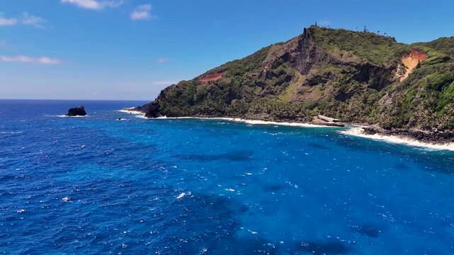 Aerial Drone footage of Pitcairn Islands Over Clear Blue Ocean. Volcanic coast ocean showcasing rugged green slopes, basalt cliffs, scattered village houses, crashing white waves over coral