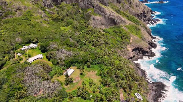 Aerial Drone footage of Pitcairn Islands Over Clear Blue Ocean. Volcanic coast ocean showcasing rugged green slopes, basalt cliffs, scattered village houses, crashing white waves over coral