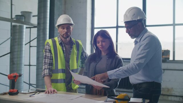 Engineer and contractor in hard hats explaining construction plan to female client and showing her around building site
