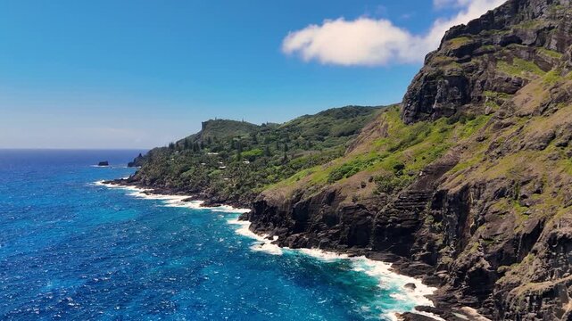 Aerial Drone footage of Pitcairn Islands Over Clear Blue Ocean. Volcanic coast ocean showcasing rugged green slopes, basalt cliffs, scattered village houses, crashing white waves over coral