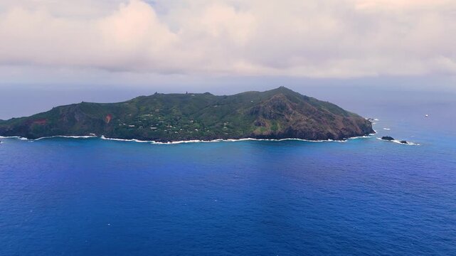 Aerial Drone footage of Pitcairn Islands Over Clear Blue Ocean. Volcanic coast ocean showcasing rugged green slopes, basalt cliffs, scattered village houses, crashing white waves over coral