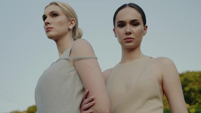 Two confident young women in elegant dresses, posing outdoors with a serene, natural background. Their poised expressions and modern style embody grace and sophistication.