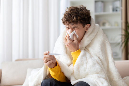 Sick teen boy wrapped in white blanket blowing nose with tissue holding thermometer on sofa. Young male with flu fever and cold symptoms at home