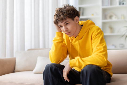 Sad bored teen boy in yellow hoodie sitting alone on sofa with hand on cheek, looking down. Lonely depressed young male feeling upset at home