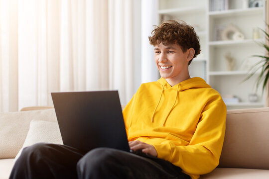 Happy young teen in yellow hoodie sitting on sofa with laptop, smiling and looking away. Cheerful male student using computer at home casually