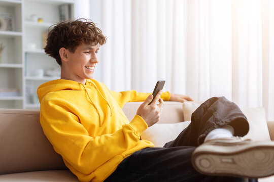 Smiling young teen male in yellow hoodie sitting on couch using smartphone. Casual relaxed guy browsing phone at home with feet up on sofa