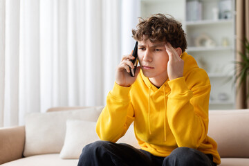 Obraz premium Worried teen boy in yellow hoodie talking on phone with hand on temple, looking stressed. Young troubled male having difficult call at home on sofa