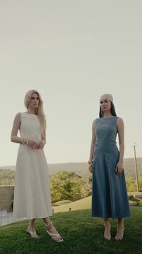 Two elegant women dressed in stylish garments, posing outdoors on a bright sunny day with scenic greenery in the background. Elegant Women in Stylish Dresses Standing Outdoors on a Sunny Day