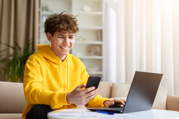 Fototapeta premium Happy teen boy in yellow hoodie using smartphone and laptop simultaneously at home, smiling. Young male multitasking with phone and computer on sofa indoors