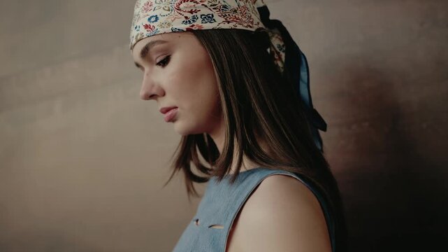 A thoughtful young woman wearing a floral bandana and blue top, looking down pensively in front of a neutral background with subdued lighting. Young Woman Wearing Floral Headscarf