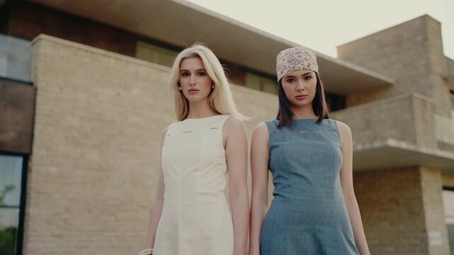 Two fashionable women posing in stylish dresses, one in white and the other in denim, standing confidently outside a modern building. A scene depicting contemporary fashion and confidence.