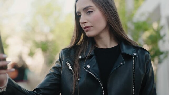 Young woman taking a selfie outdoors on a sunny day, wearing a stylish leather jacket. Capturing a confident and modern lifestyle moment. Young Woman Taking Selfie Outdoors in Leather Jacket