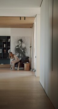 A woman in pajamas walks through her modern apartment and sits in a comfortable chair. The apartment has a minimalist design with wooden floors and neutral colors.
