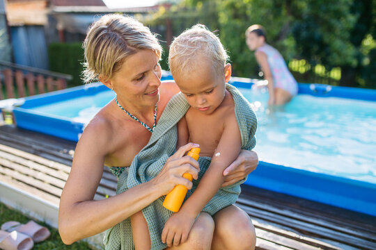 Mother applying sunscreen to toddler child near backyard pool on sunny summer day.