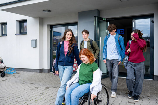 Group of teenage students supporting friend in wheelchair in front of school building.