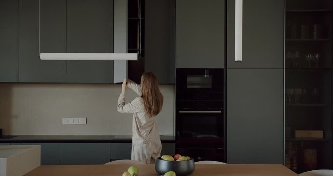 Woman in pajamas opens cupboard in modern kitchen. The contemporary design features dark gray cabinets and sleek appliances. She reaches for something inside, showcasing the practicality and style