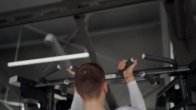 Focused young man in a white shirt utilizing gym equipment for his workout, shot from behind in a modern gym setting. Young Man Engaged in Intense Workout on Gym Equipment