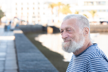 Elderly bearded man smiling in profile on a sunny seaside promenade wearing a striped shirt © simona