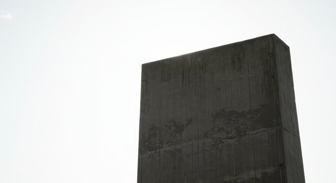 A colossal, textured concrete block silhouette dominates the frame, standing starkly against a bright, minimalist white sky backdrop, rigidity, massive, foundation