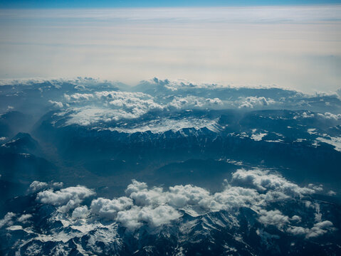 Aerial view of the Alps mountains from airplane window above clouds. Snow-covered peaks, scenic landscape, travel and aviation concept in Europe.