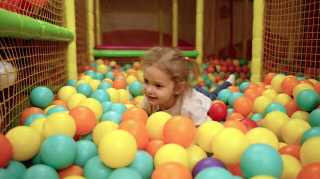 Playground with ball pit indoor. Joyful smiling cute little girl having fun at indoor play center, playing with ball. Slow motion