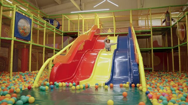 Happy joyful little girl playing at indoor play center playground, sliding in colored slide into balls in ball pool. Slow motion