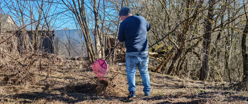 Man wearing jeans and a blue sweater raking dry leaves and branches in a neglected backyard during a sunny early spring day, engaging in garden cleanup and yard work.