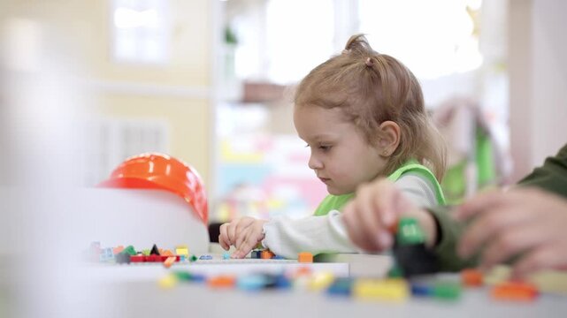 Cute little Caucasian girl playing with friend while playing with colorful toy blocks on the table in educational gaming center. Slow motion