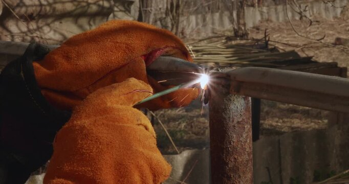 Close-up of a worker in protective gloves using an electrode to weld a rusty metal pipe, creating bright sparks and smoke during the arc welding process for fence construction and repair.