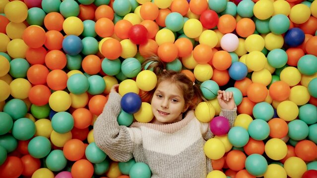 Happy little joyful cute girl playing with colorful balls in playground ball pool, lying on the floor in game center. Slow motion. Childhood, entertainment, amusement park concept