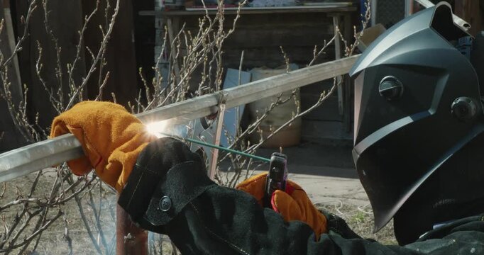 Professional welder in a protective mask and gloves performs welding work on a metal structure, creating bright sparks and smoke while connecting steel parts with an electrode outdoors.