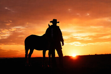 Fototapeta premium A cowboy and horse stand silhouetted against a vibrant sunset over a prairie landscape.