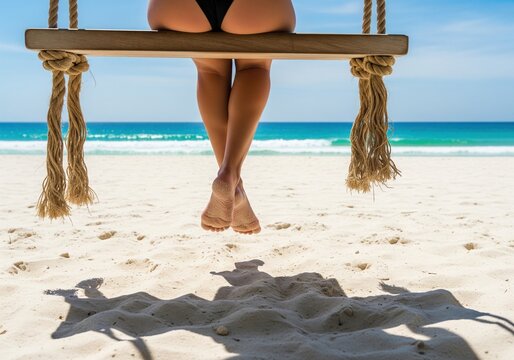 Woman in bikini sitting on wooden rope swing on sandy beach against clear blue ocean water