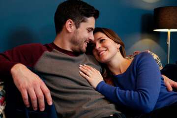 Obraz premium Caucasian young adult couple cuddling on sofa, smiling and leaning together, expressing intimacy and relationship trust. Ideal for dating, marriage, home life, or Valentine marketing