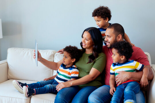 Wide shot of multiethnic family using tablet while sitting together on sofa, sharing video call or selfie moment. Supporting parenting, family connection, home technology marketing