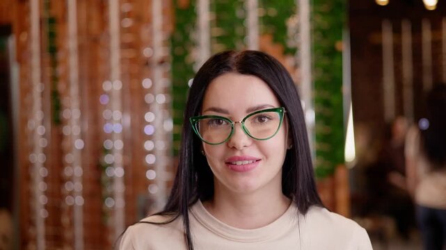 Static shot of woman wearing green glasses smiling and nodding in indoor cafe with bokeh string lights and shallow depth of field.